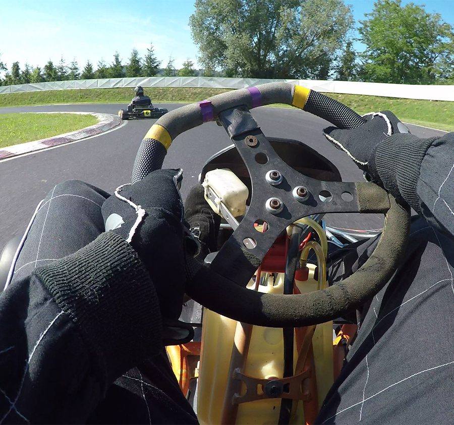 POV: Steering a speeding go cart along the winding asphalt circuit on a sunny autumn day. Driving a fast cart in a black suit through sharp turns of a bumpy racetrack. Bright sun shines on racers